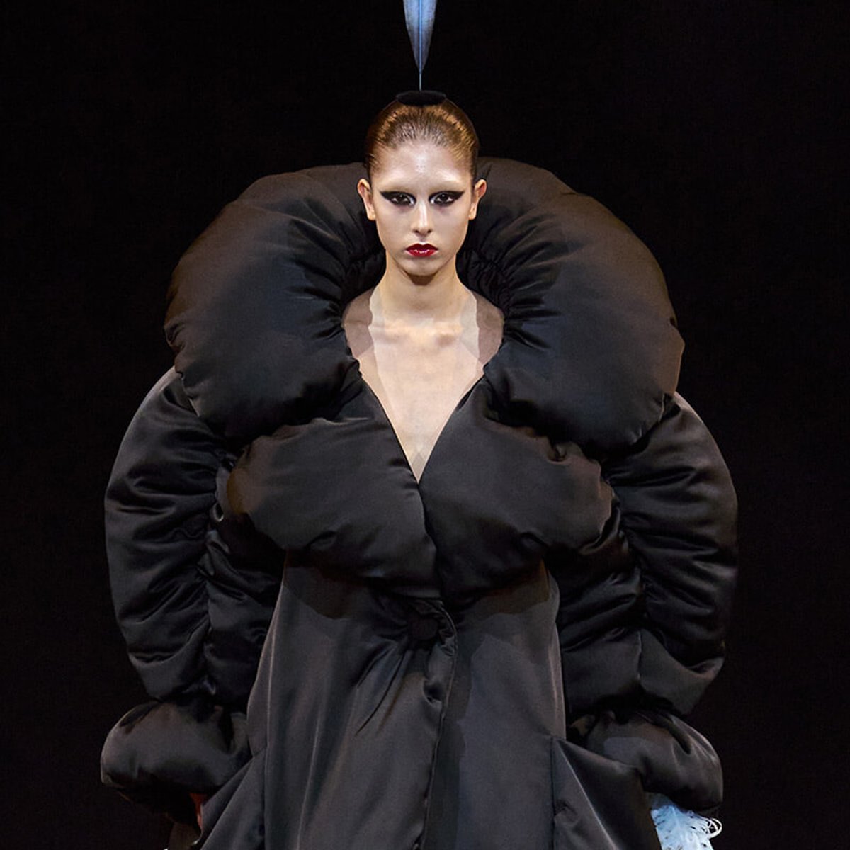 Runway model in an oversized black padded coat with huge rounded shoulders on a dark stage.
