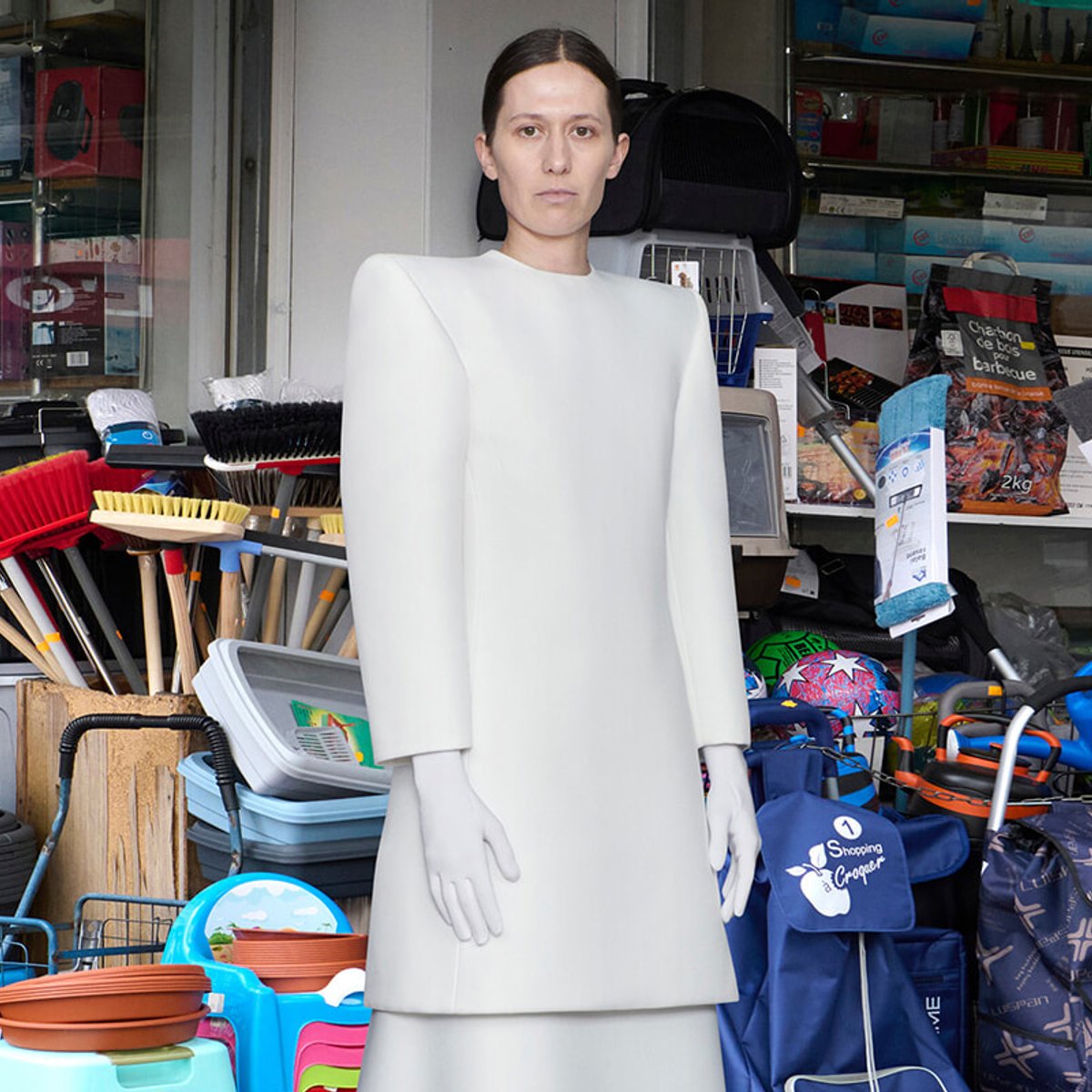 Person wearing a sculptural white dress stands outside a cluttered shop, with brooms, bags, and colorful containers in the background.