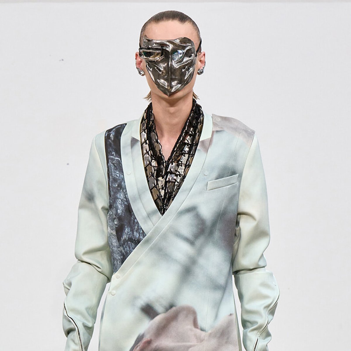 Model on a runway wearing a futuristic metallic mask and a pale satin wrap blazer with a dark, net-like inner layer.
