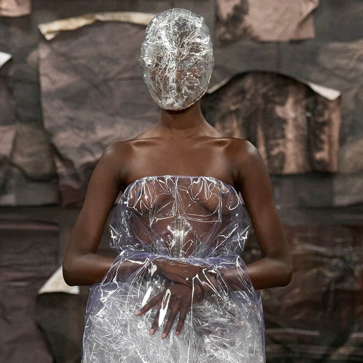 Model wearing a transparent plastic wrap gown and matching headwrap in a dark, textured backdrop of rocks or fabric.
