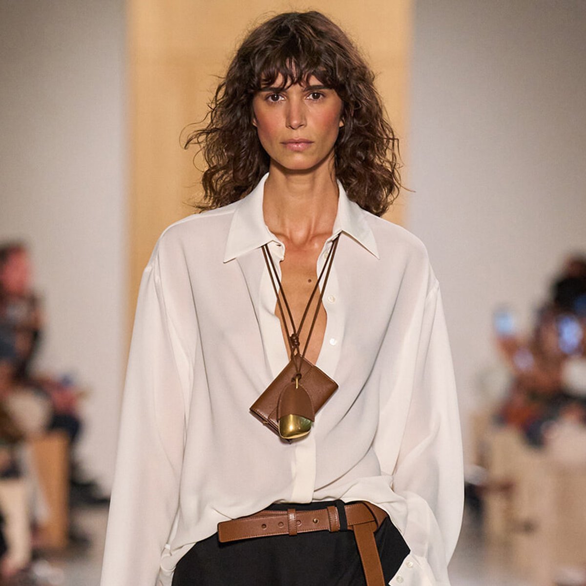 Model walking a fashion runway in a white oversized shirt, brown belt, and layered leather necklace with a small pendant bag accent.