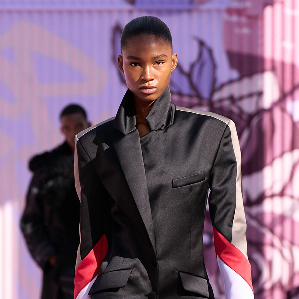 Fashion model walks the runway in a black blazer with bold red and white color-block sleeves, on a purple-toned backdrop.