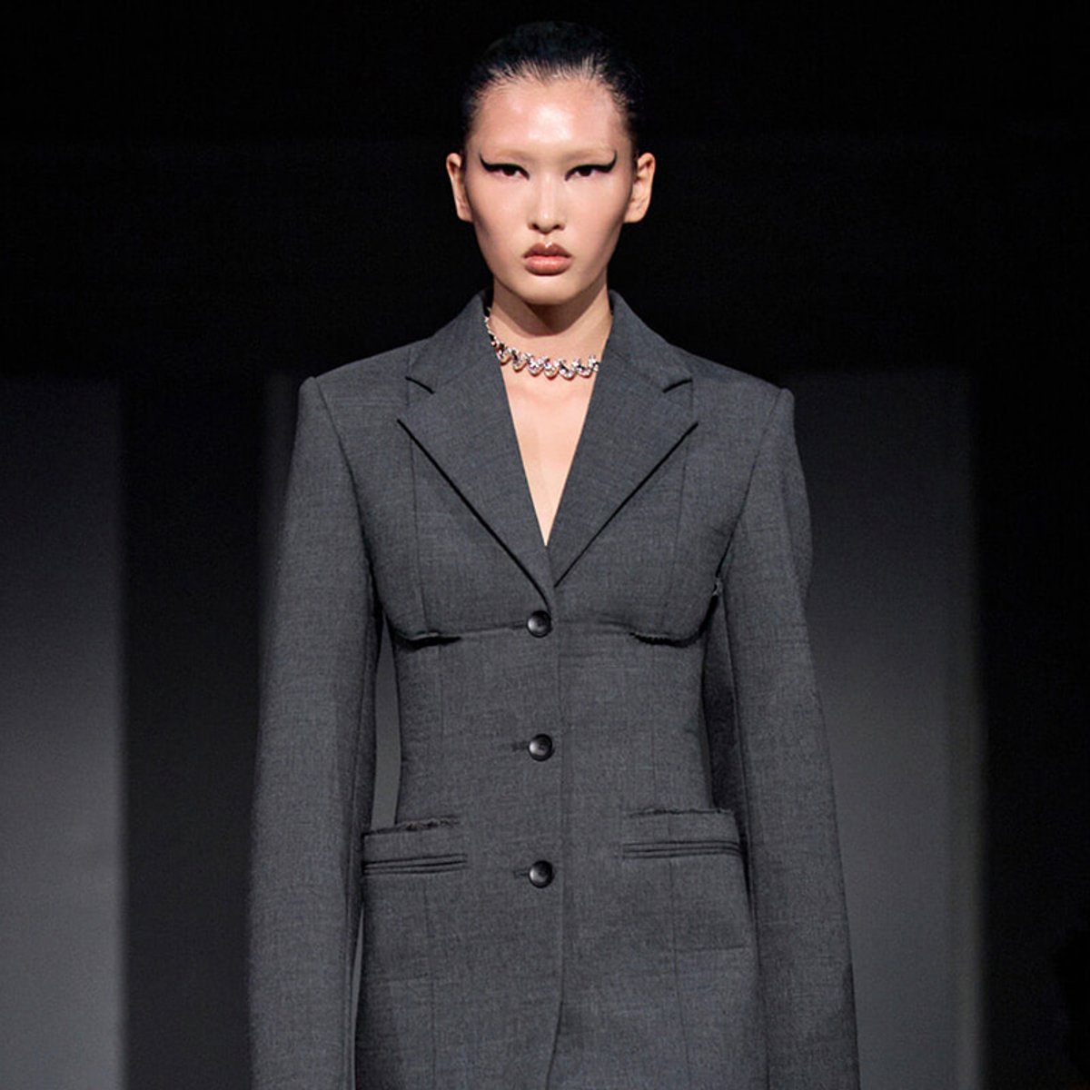 Female model wearing a dark grey tailored blazer with a notched lapel and matching trousers on a fashion runway.