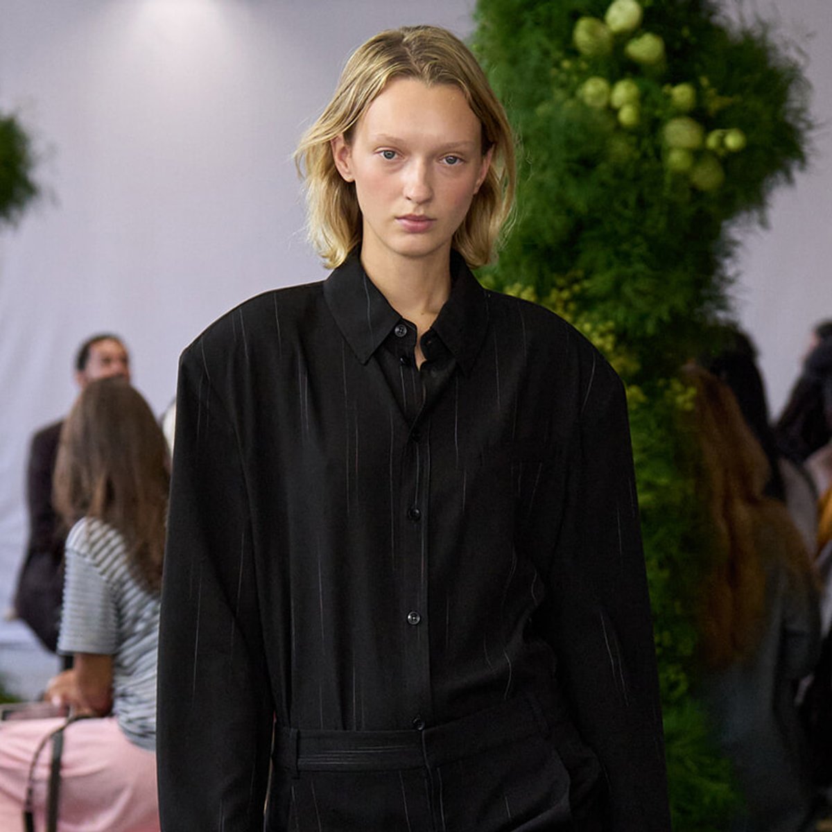 Model walks a runway in a loose black pinstriped shirt with a belt, serious expression, backstage setting with greenery in the background