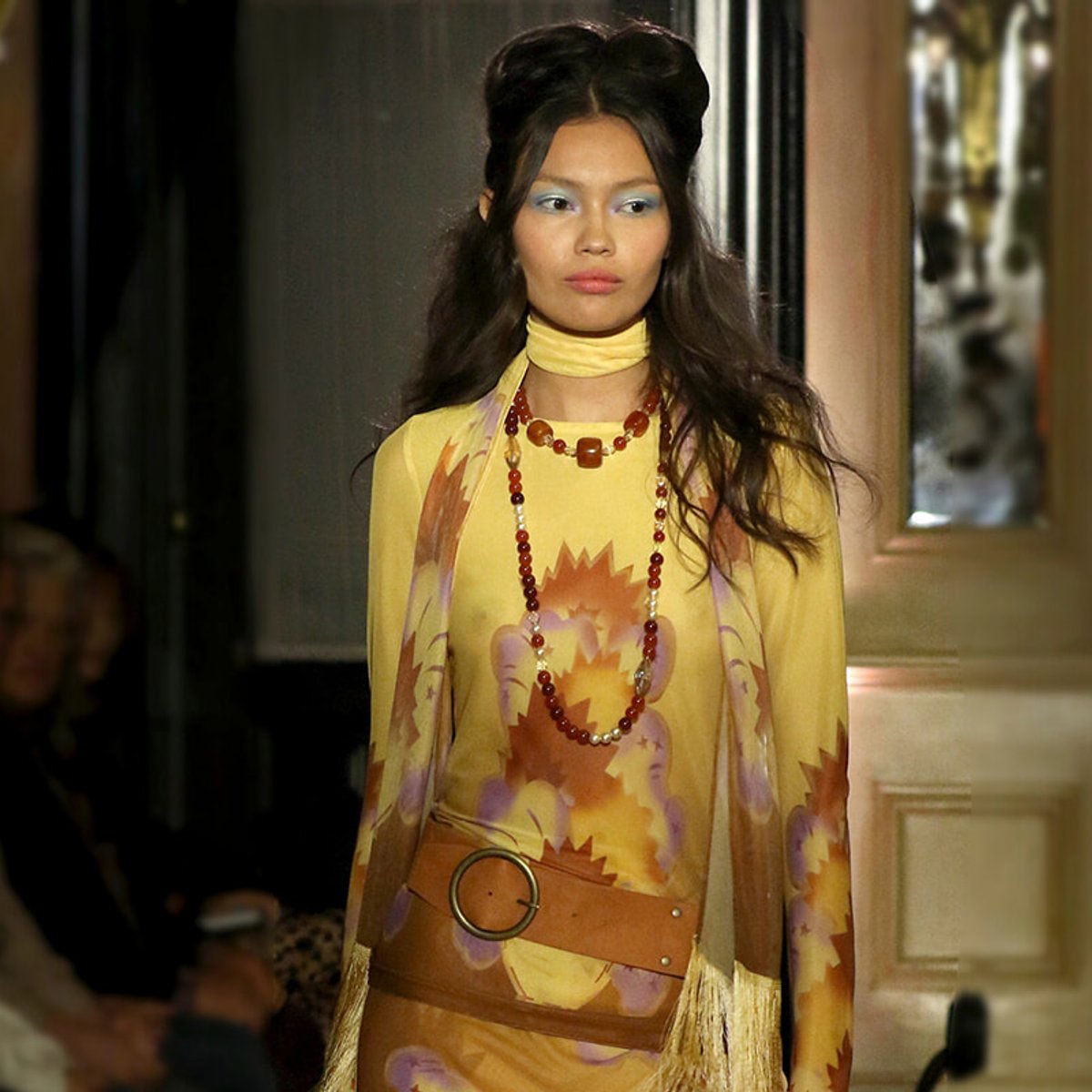 Model walking a runway in a yellow, long-sleeve dress with a bold orange and purple print, layered red necklace, and tan belt with fringe.
