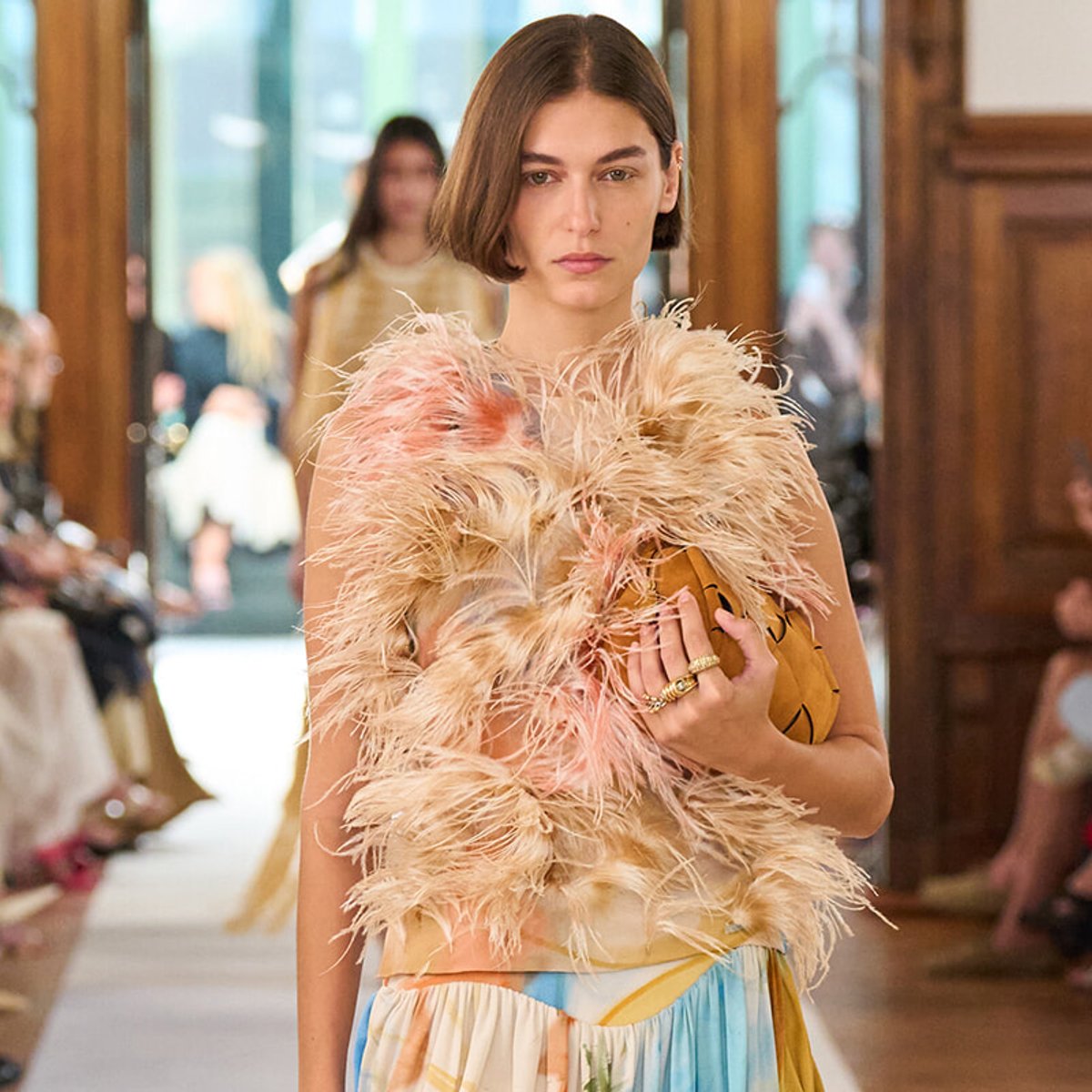 Model walking a runway in a feathered pink-beige top, holding a tan clutch amid a fashion show crowd.