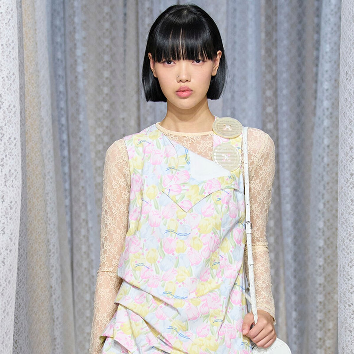 Young Asian female model walks the runway in a pastel floral dress with lace sleeves and a white shoulder bag.