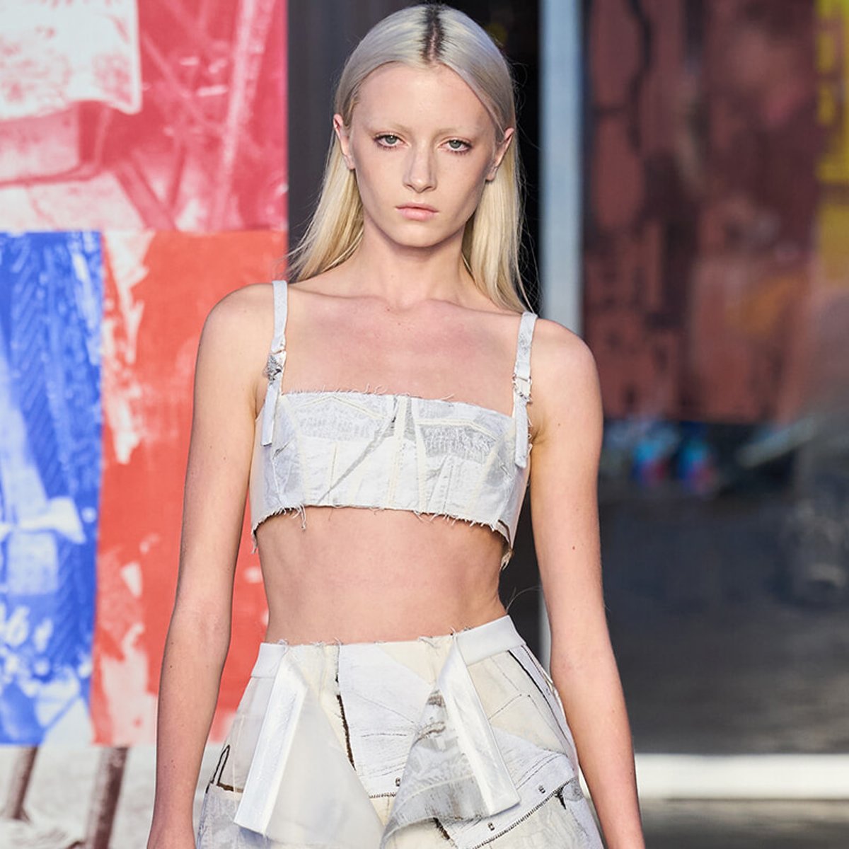 Female model walking a runway in a white textured crop top and matching high-waisted skirt with frayed edges.