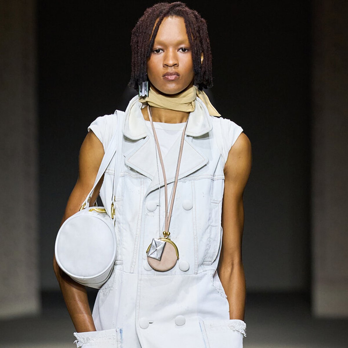 Model walking a runway in a white sleeveless vest with a beige scarf and a round white bag slung over the shoulder.