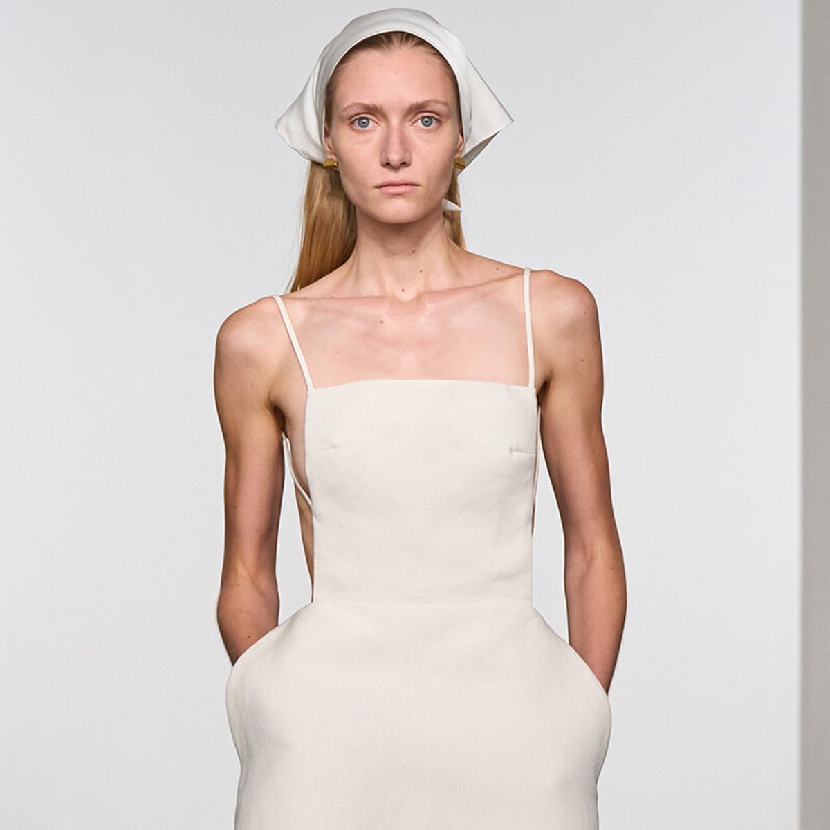 Model walking a fashion show in a cream spaghetti-strap dress with a white headscarf against a neutral background.