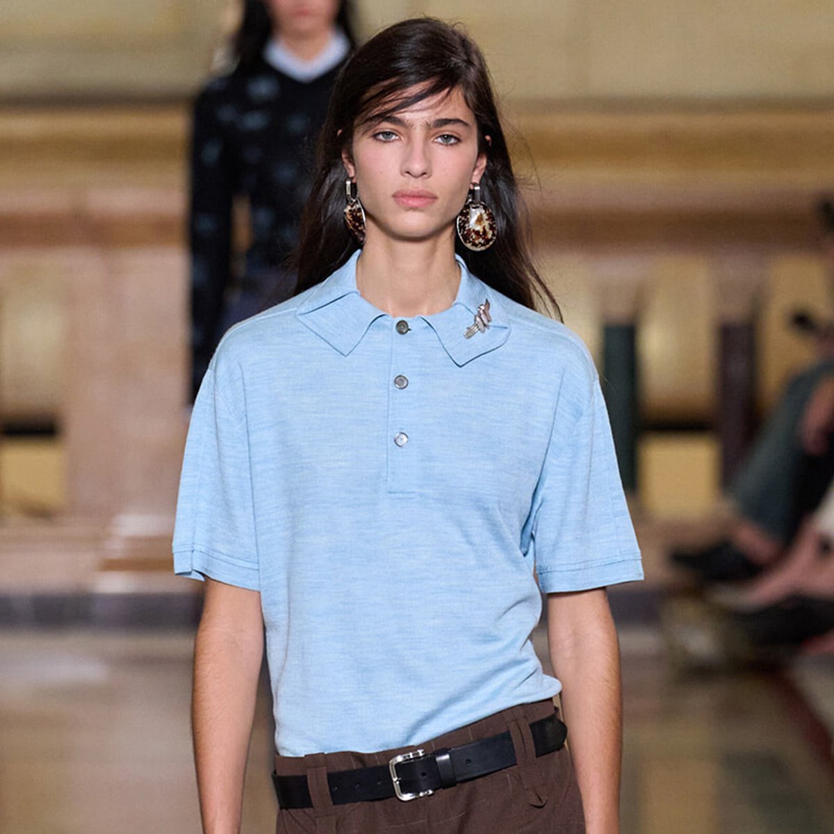 Female model walking the runway in a light blue polo shirt with a pointed collar, brown belt and large dangling earrings; blurred fashion-show backdrop behind her.