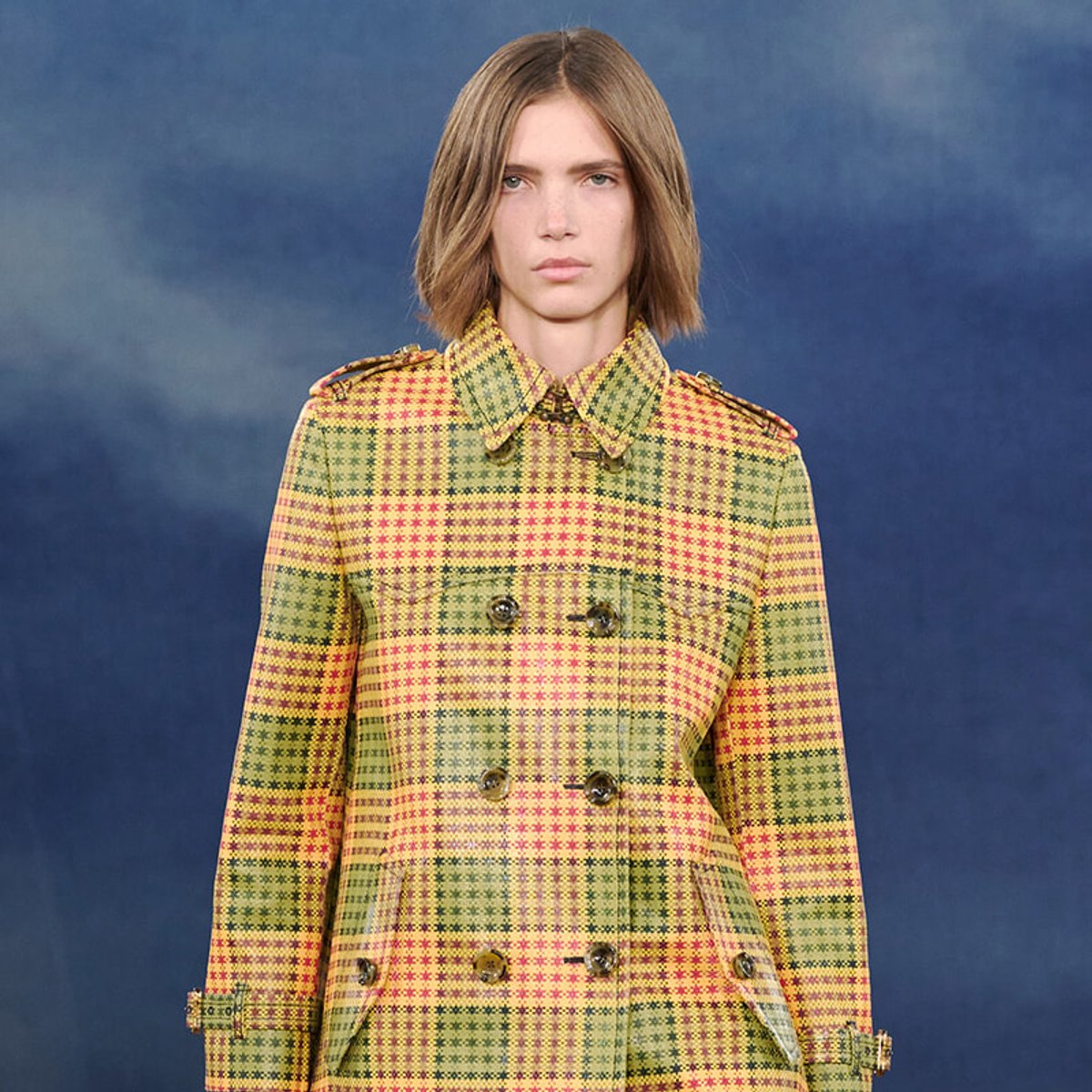 Model wearing a bright yellow, red, and green plaid double-breasted coat against a blue backdrop.