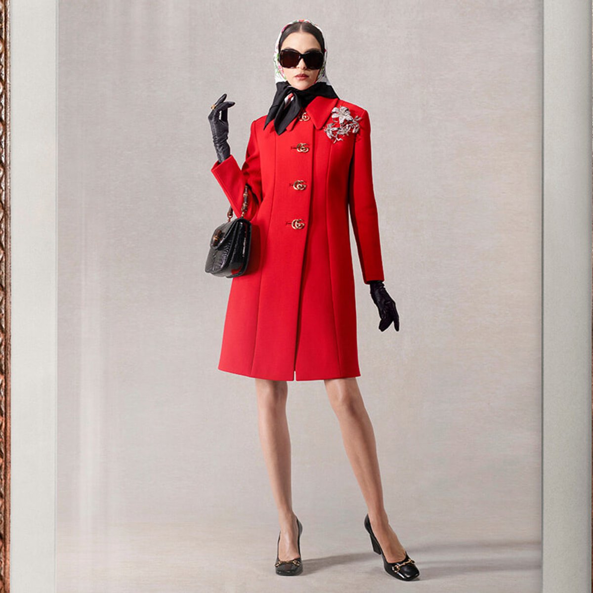 Woman in a bright red knee-length coat with gold buttons, black gloves, and a patterned headscarf, posing with a black handbag and sunglasses.