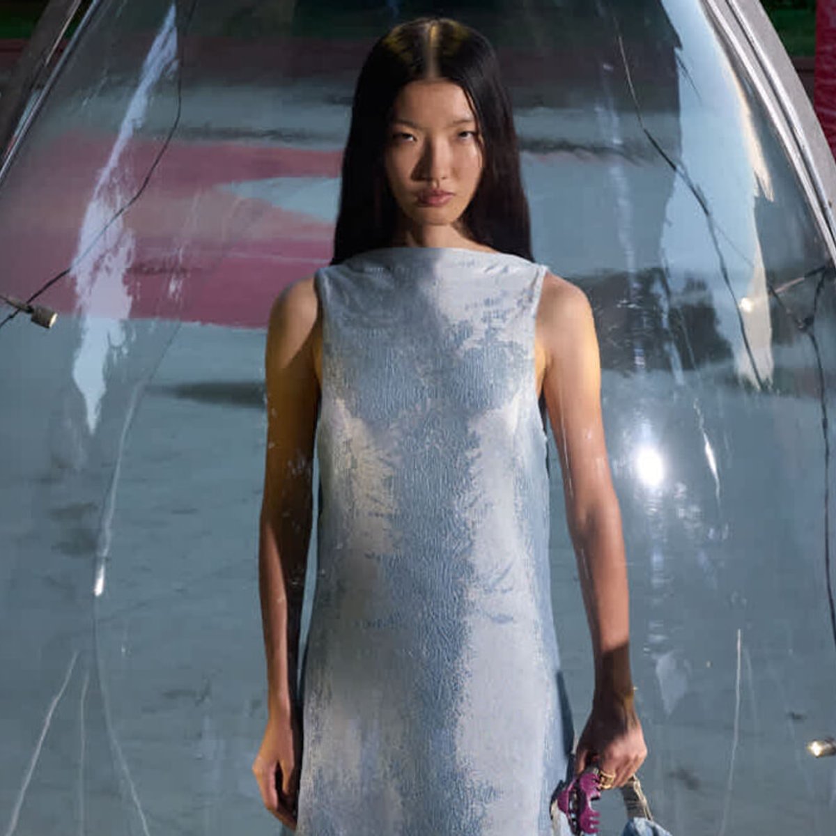 Young woman with long black hair wearing a pale blue sleeveless dress, standing in front of a large reflective windshield, holding a small pink accessory.