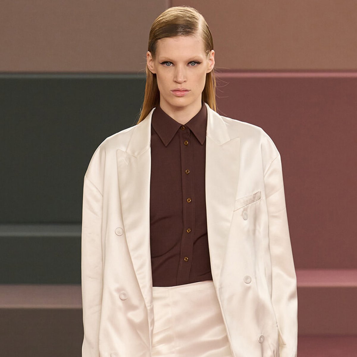 Model walking a runway wearing a cream oversized blazer over a dark brown button-up shirt, neutral makeup, and slicked-back hair.