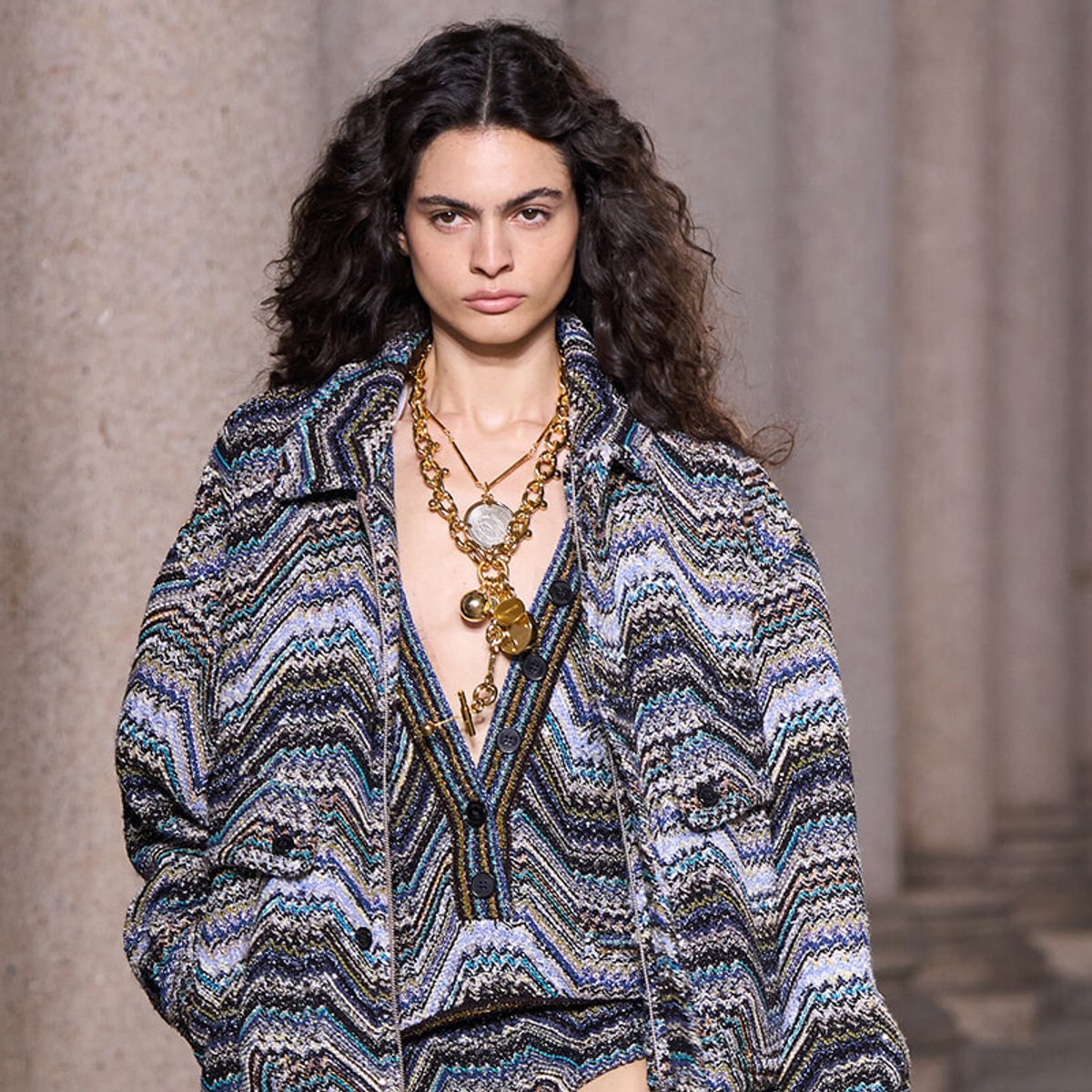 Model on a runway wearing a blue-gray zigzag patterned coat with layered chunky gold necklaces.