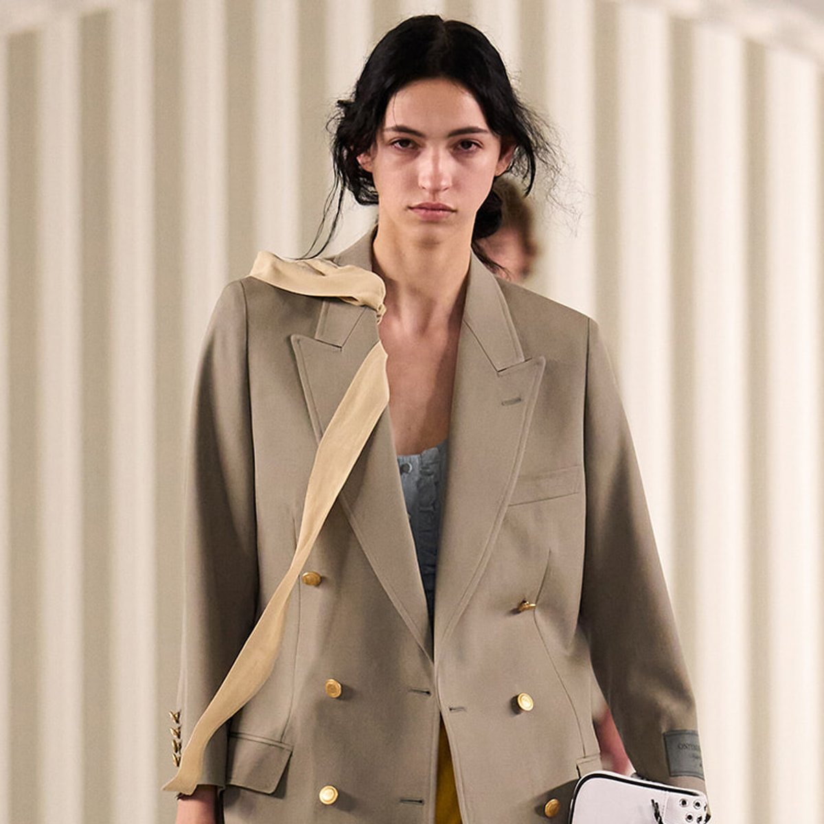 Female model on a runway wearing an oversized beige double-breasted blazer with gold buttons and a cream scarf draped over one shoulder.