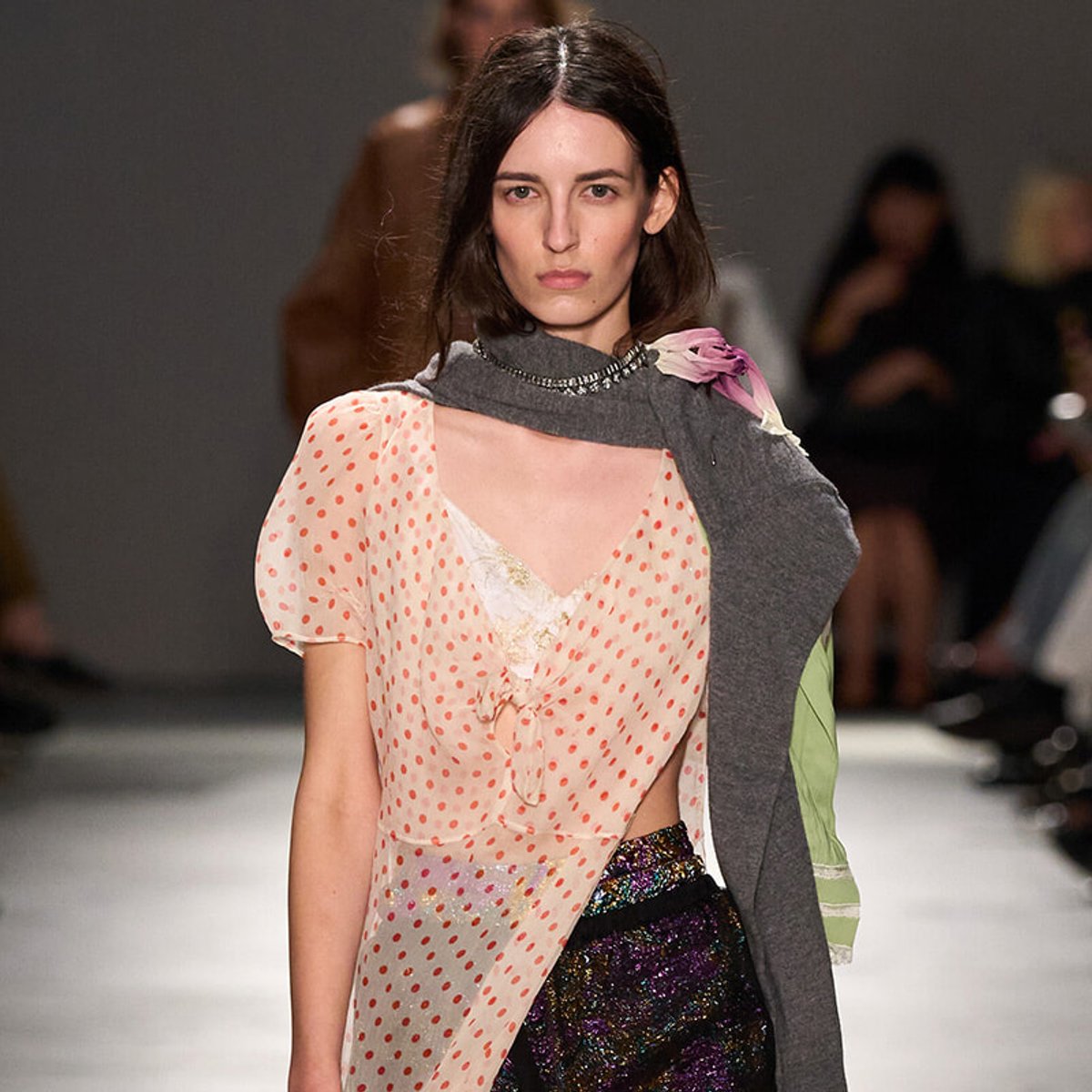 Model walking a runway wearing a pink polka-dot sheer blouse with lace trim and a gray scarf draped over the shoulder.
