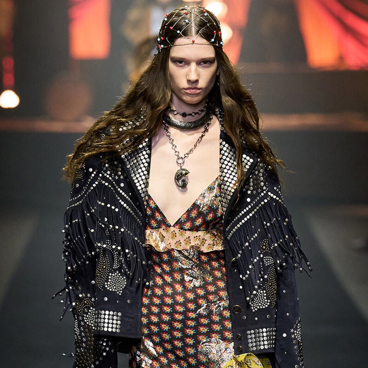 Model walking a runway in a black studded jacket with fringe over a floral dress, accessorized with layered necklaces and a beaded headpiece.