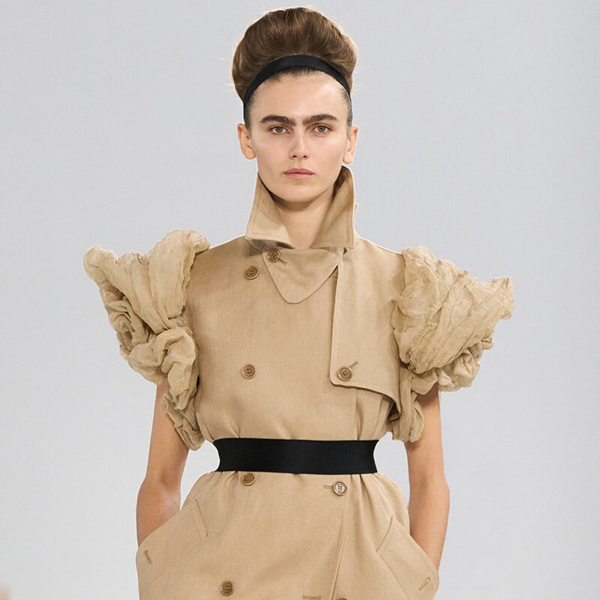 Model wearing a beige trench-inspired dress with oversized puff sleeves, a black waist belt, and a high collar on a neutral backdrop