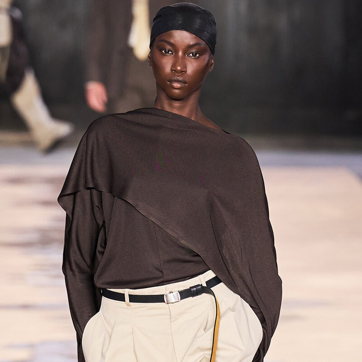 Model walking a fashion runway in a loose dark brown draped top and cream high-waisted pants with a black belt; black headscarf completes the look.