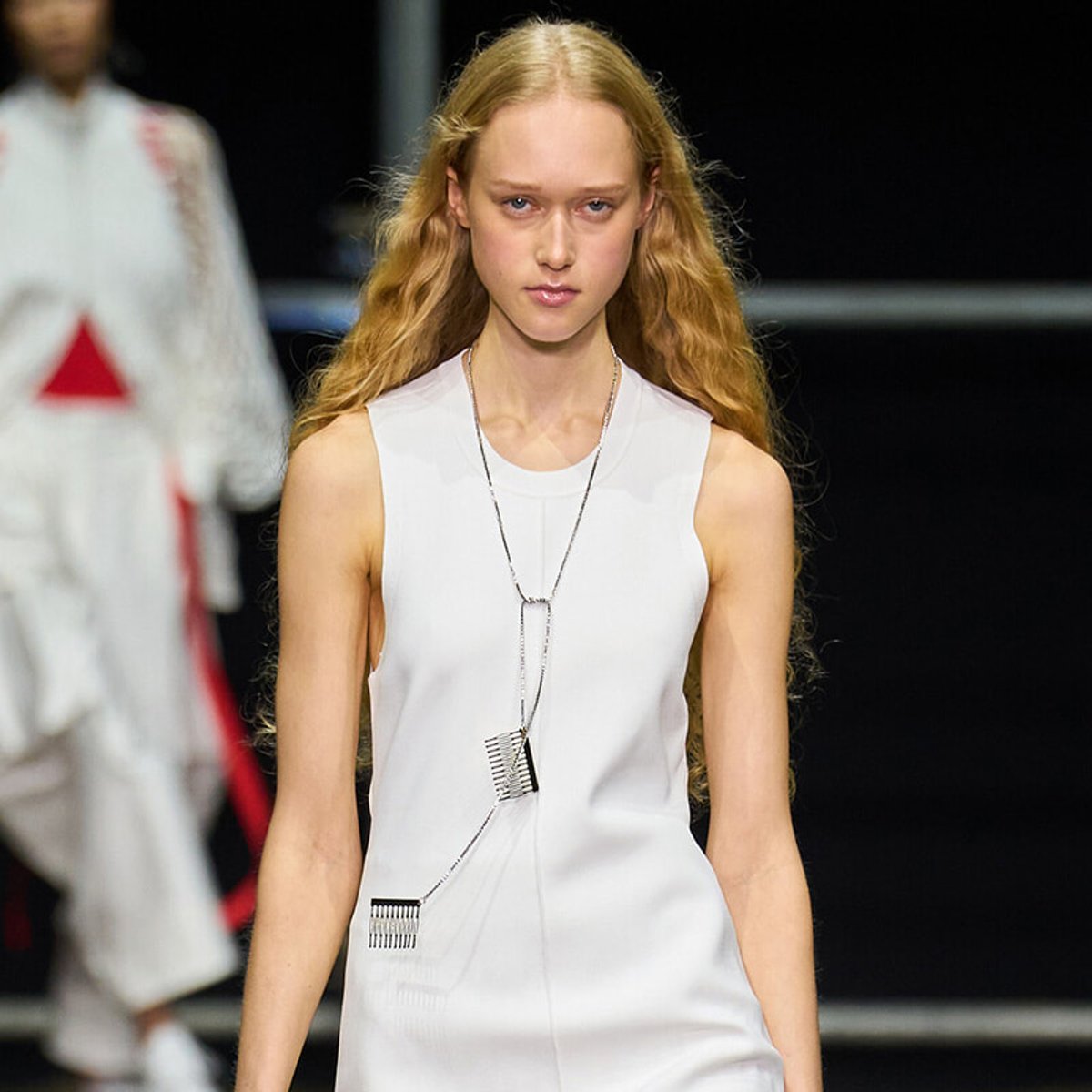 Runway model in a white sleeveless dress wearing a long geometric pendant necklace walks down the catwalk.