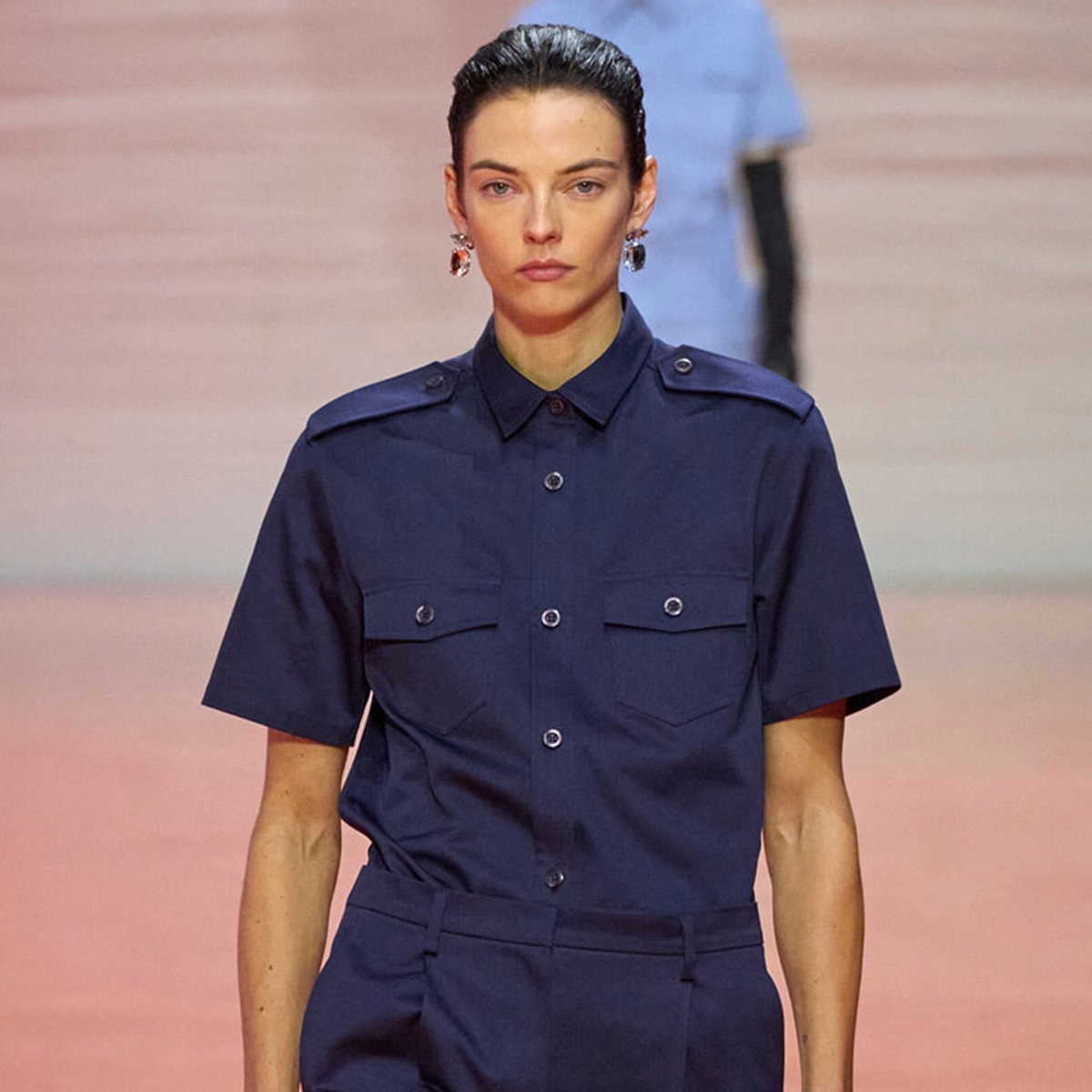 Model walks the runway wearing a navy short-sleeve utility shirt with button pockets and matching trousers.