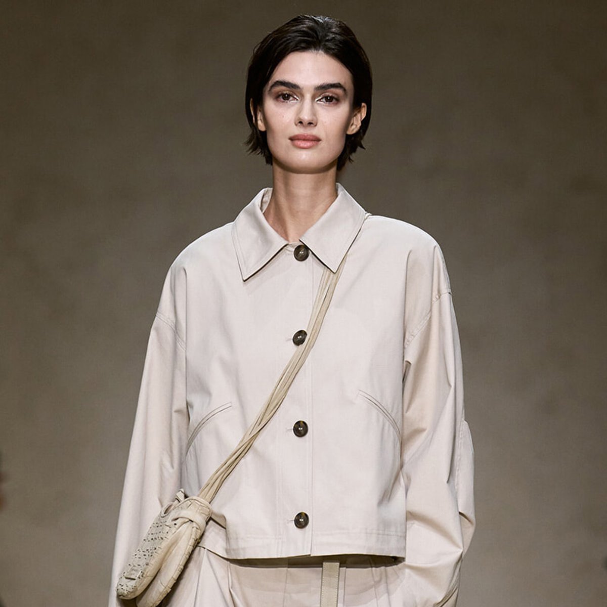 Model wearing a loose beige button-front jacket with a crossbody bag over her shoulder, neutral runway look.