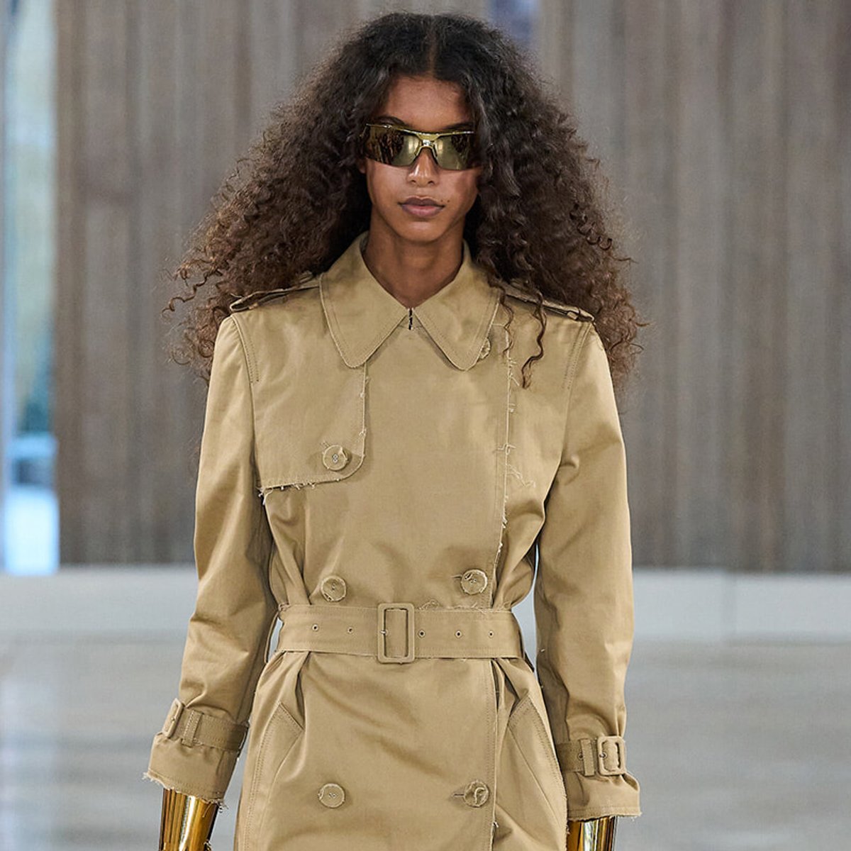 Woman model wearing a tan trench coat with belt and gold metallic gloves, sunglasses, walking on a runway.