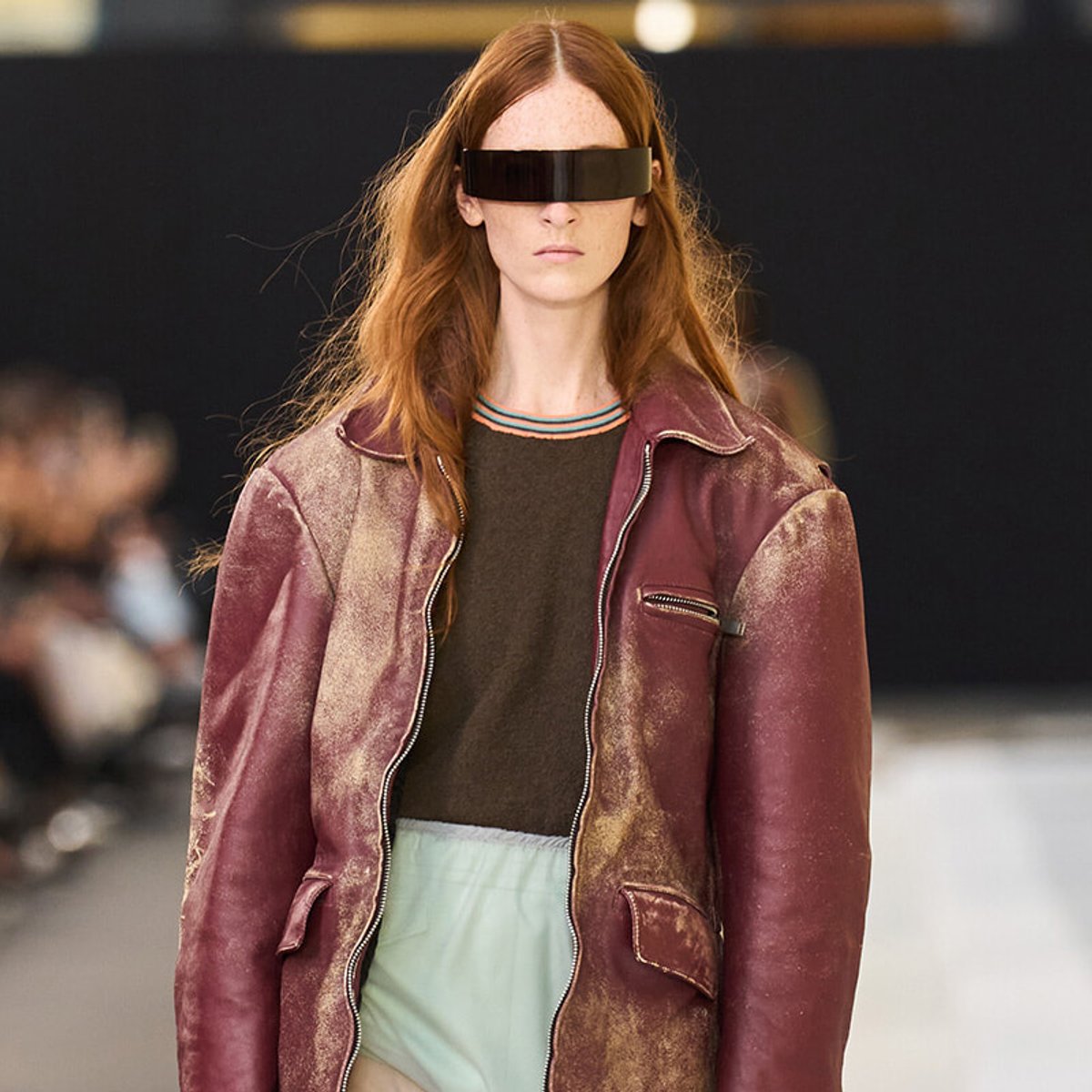 Female model on a runway wearing a maroon leather jacket, dark top, mint shorts, and a black futuristic visor over her eyes for a fashion show.