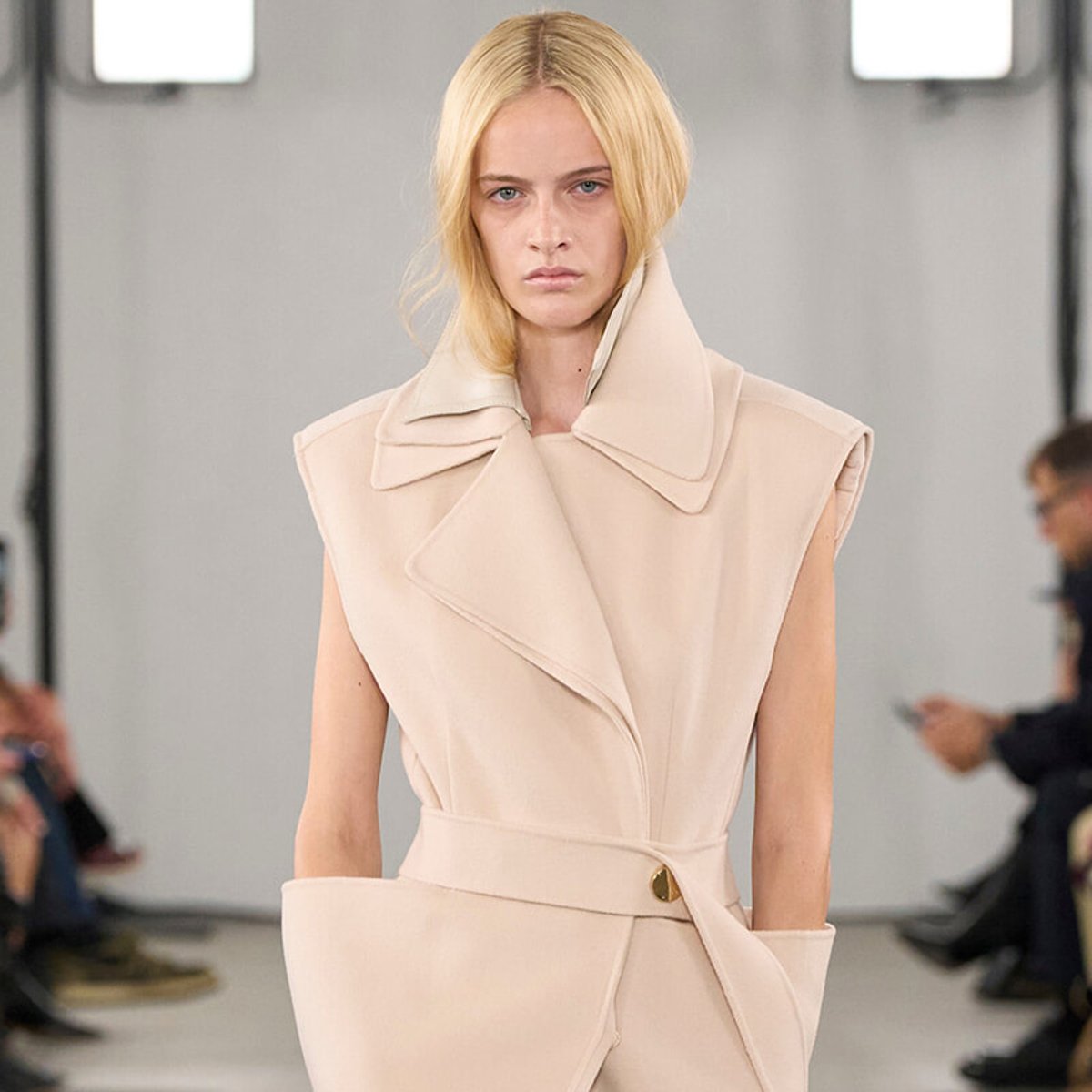 Model on a runway wearing a pale beige sleeveless coat with an oversized collar and belt.