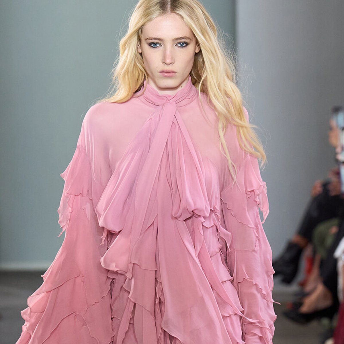 Blonde model walks runway in a pink sheer, ruffled blouse with a neck bow.