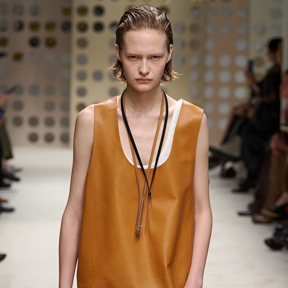 Model walking a runway in a sleeveless burnt-orange leather dress with white trim and a long black lanyard necklace.