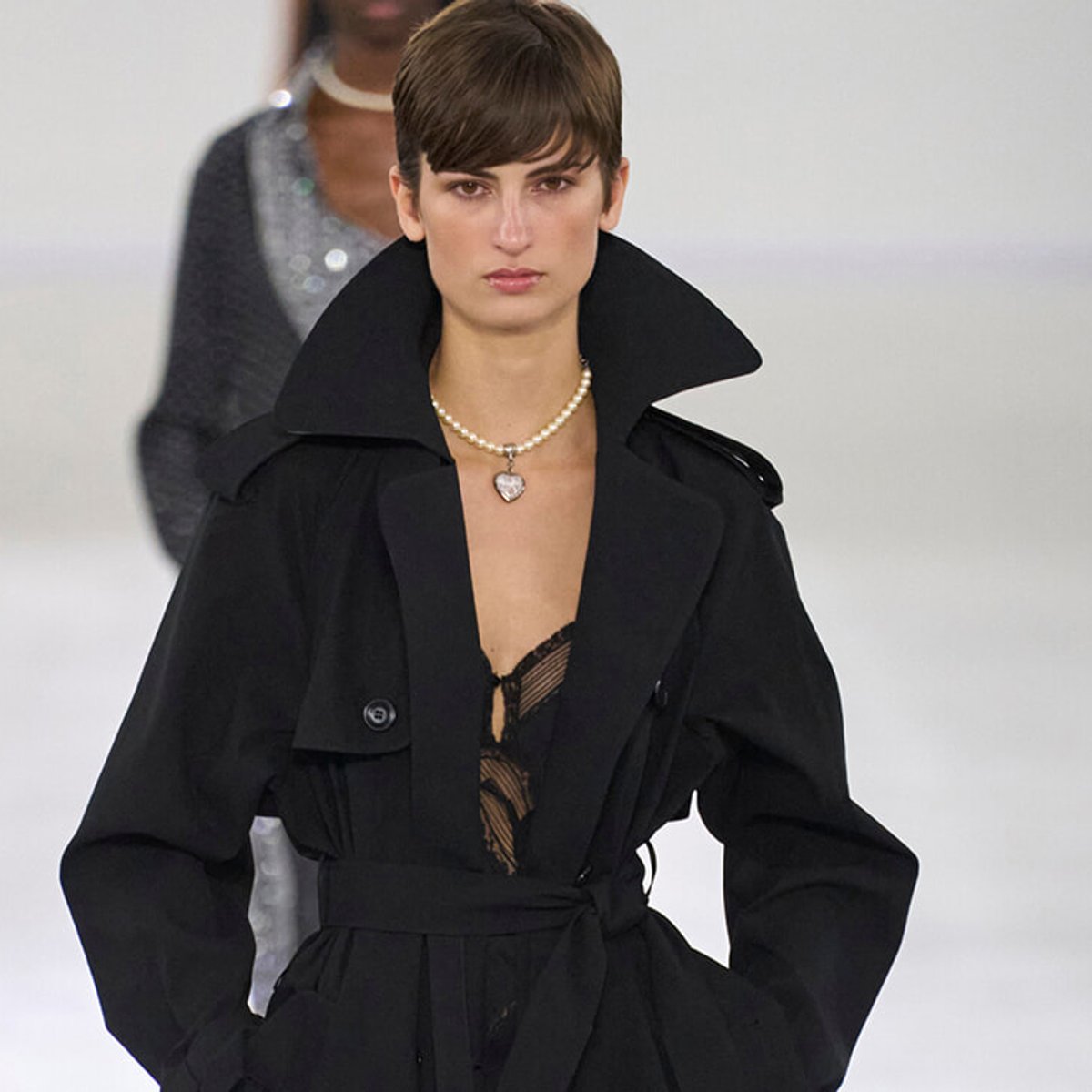 Model walking the runway in a black trench coat with an oversized collar and a pearl necklace with a heart pendant.