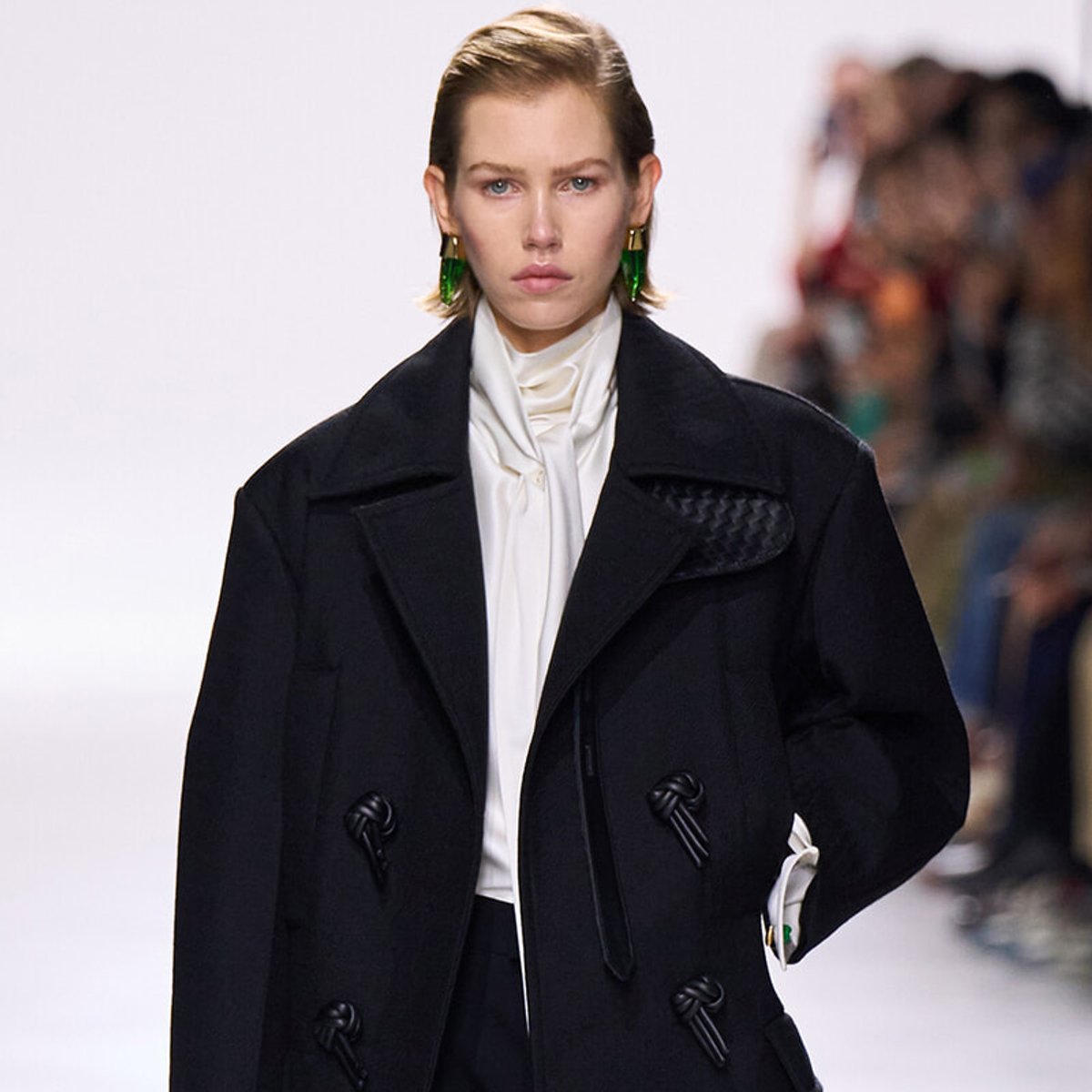Model on a runway wearing an oversized black coat over a cream blouse, with bold green earrings and a blurred audience in the background.