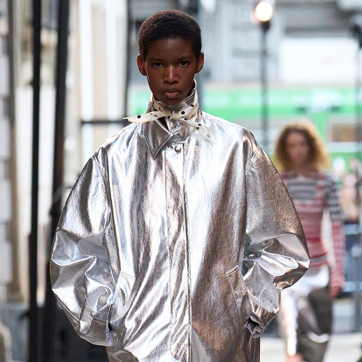 Person wearing a shiny silver cape-style coat walking on a city street, hands in pockets and focused expression