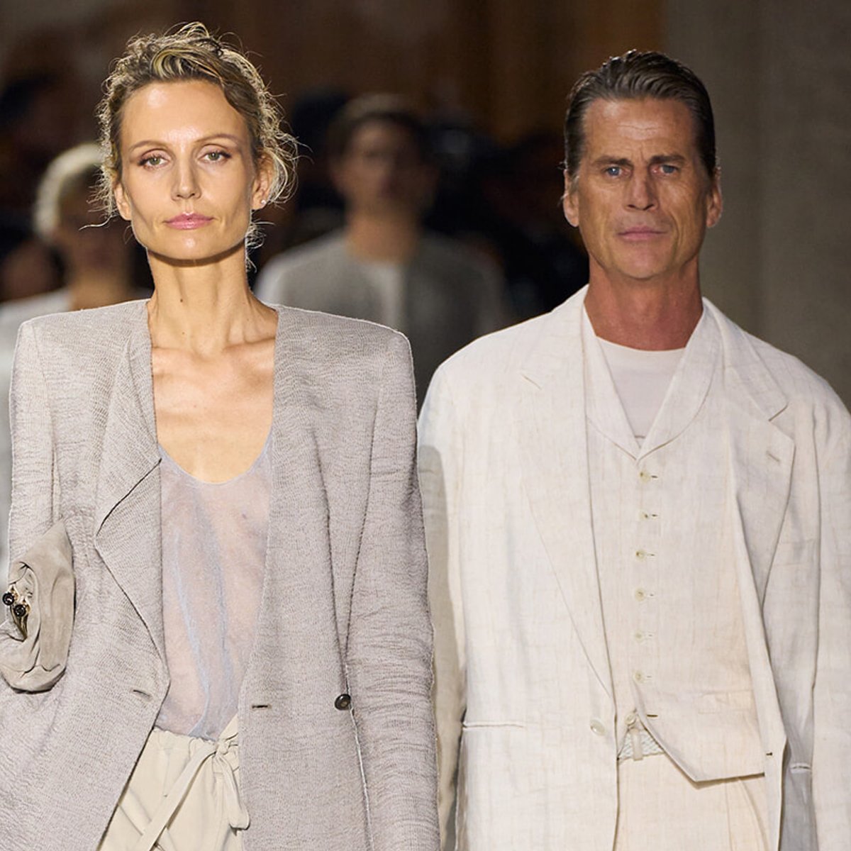Two models walk a fashion runway in neutral-toned outfits: a woman in a light blazer over a sheer top and tied-waist pants and a man in a cream suit with a vest.
