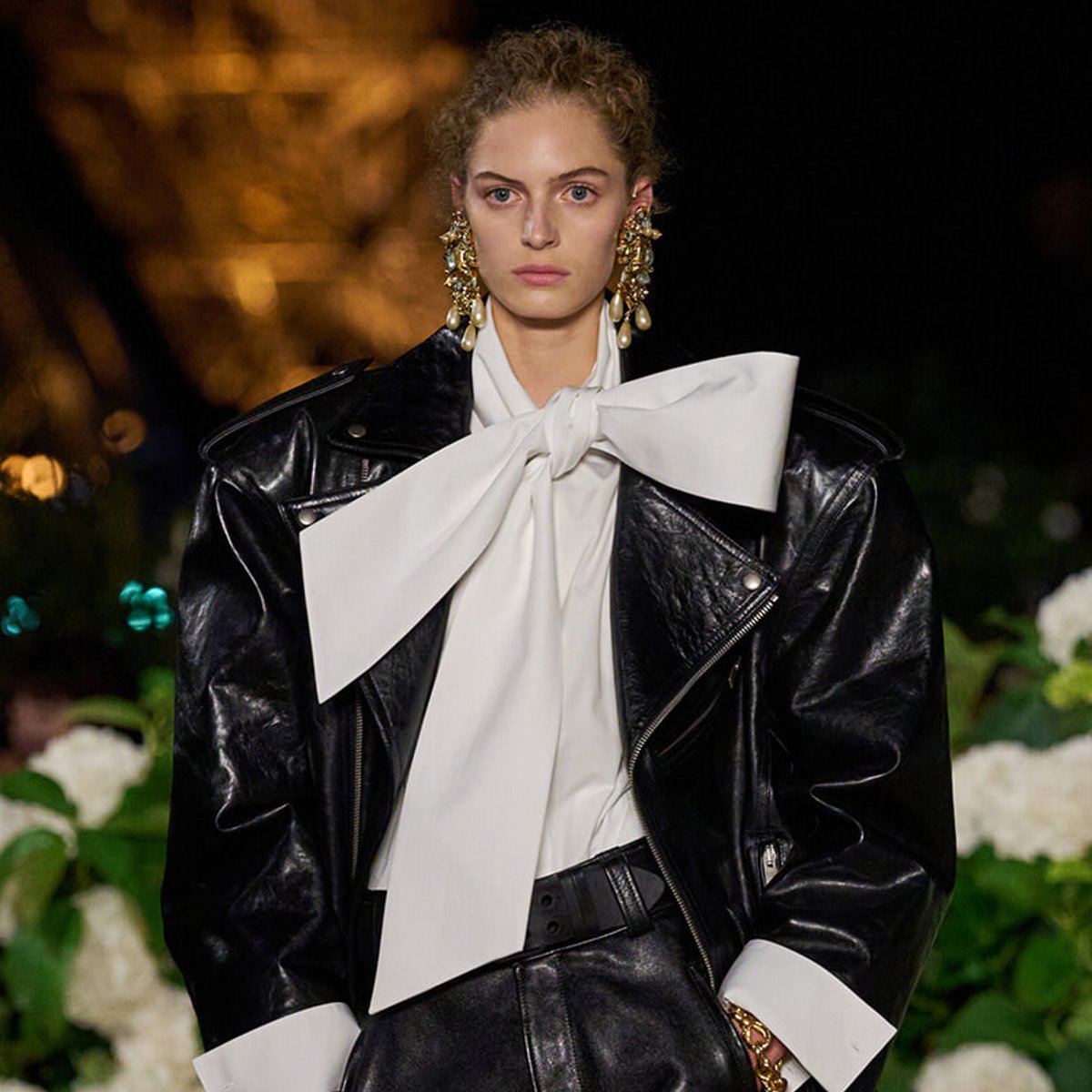 Model on a runway wearing a glossy black leather jacket with a large white bow blouse and gold chandelier earrings.