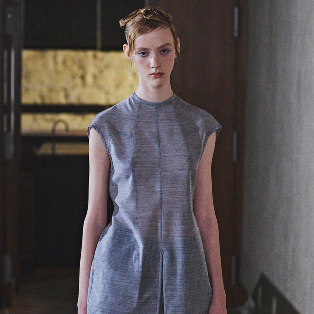 Young woman model wearing a light gray sleeveless dress walking down a backstage hallway with a neutral expression.