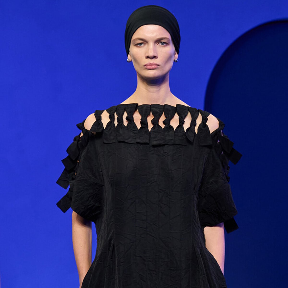 Model wearing a black off-shoulder dress with ruffled shoulder details against a blue backdrop.
