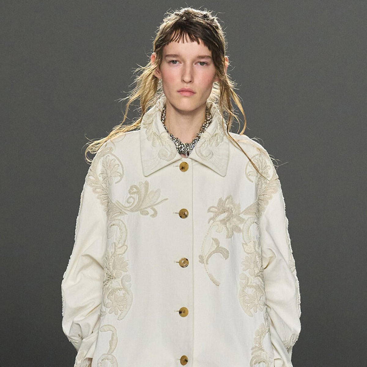 Model walks the runway in a cream embroidered coat with a high collar and wooden buttons, paired with a statement necklace.