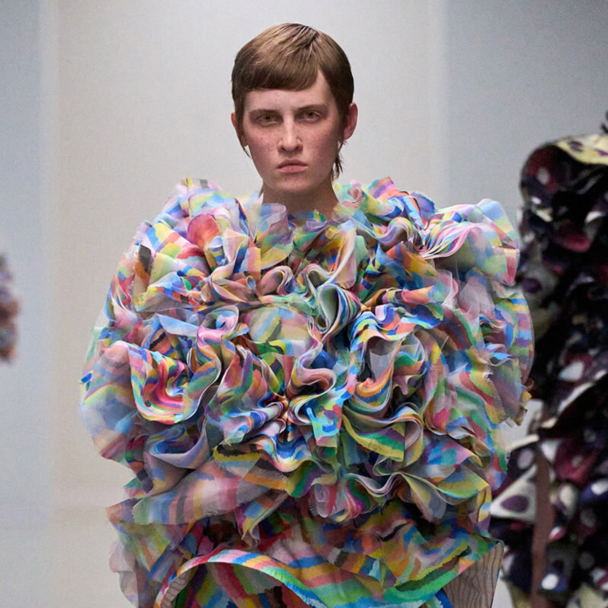 Model on a runway wearing a huge multicolored, ruffled garment with rainbow hues.