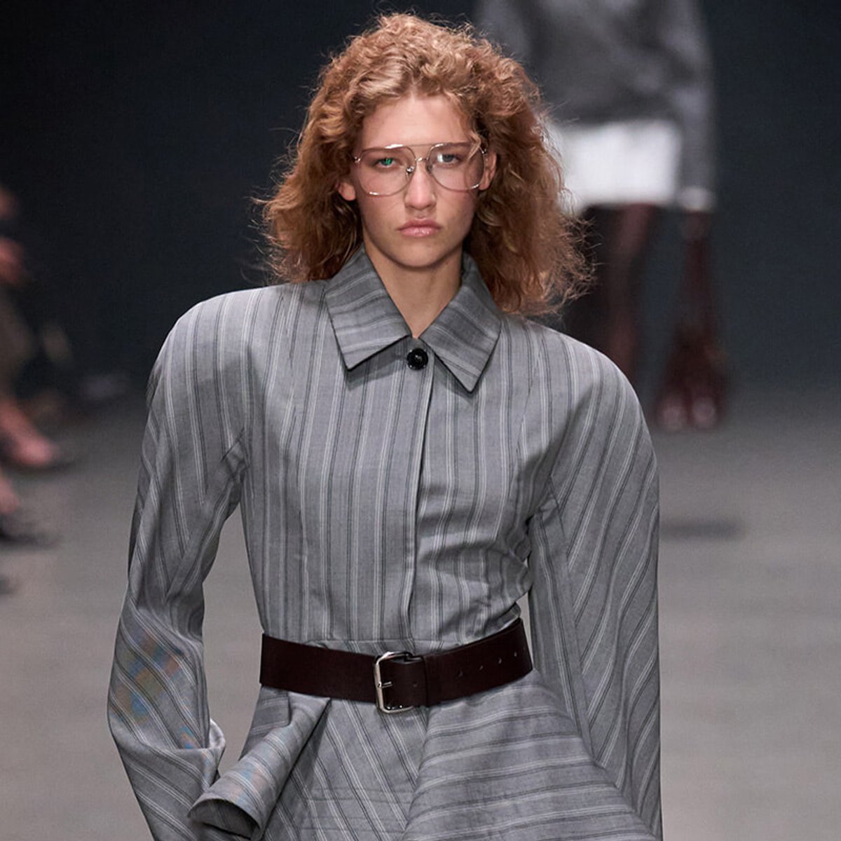 Runway model wearing an oversized gray striped shirt with a dark belt, clear glasses, and a neutral expression as she walks.
