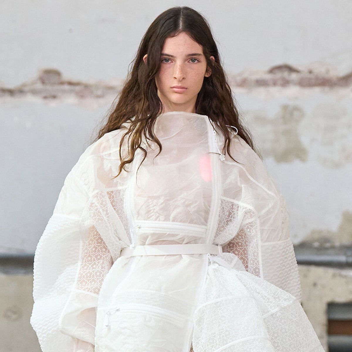 Model wearing a sheer white lace overlay dress with a belt, standing indoors.