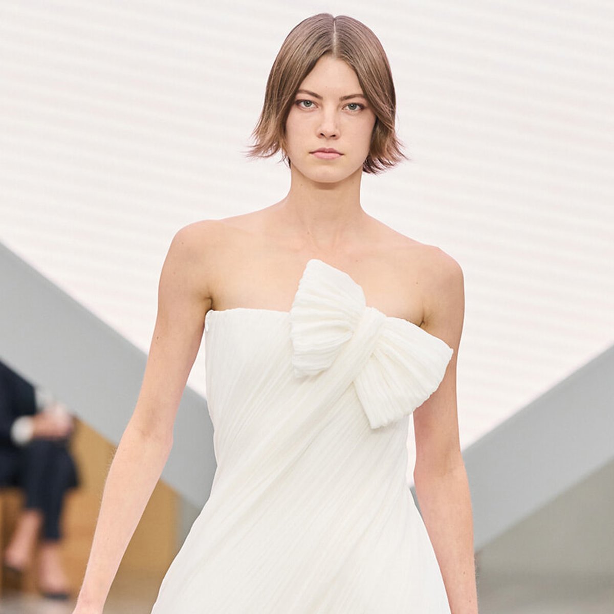 Model walking the runway in a strapless white dress with a large bow detail at the chest.