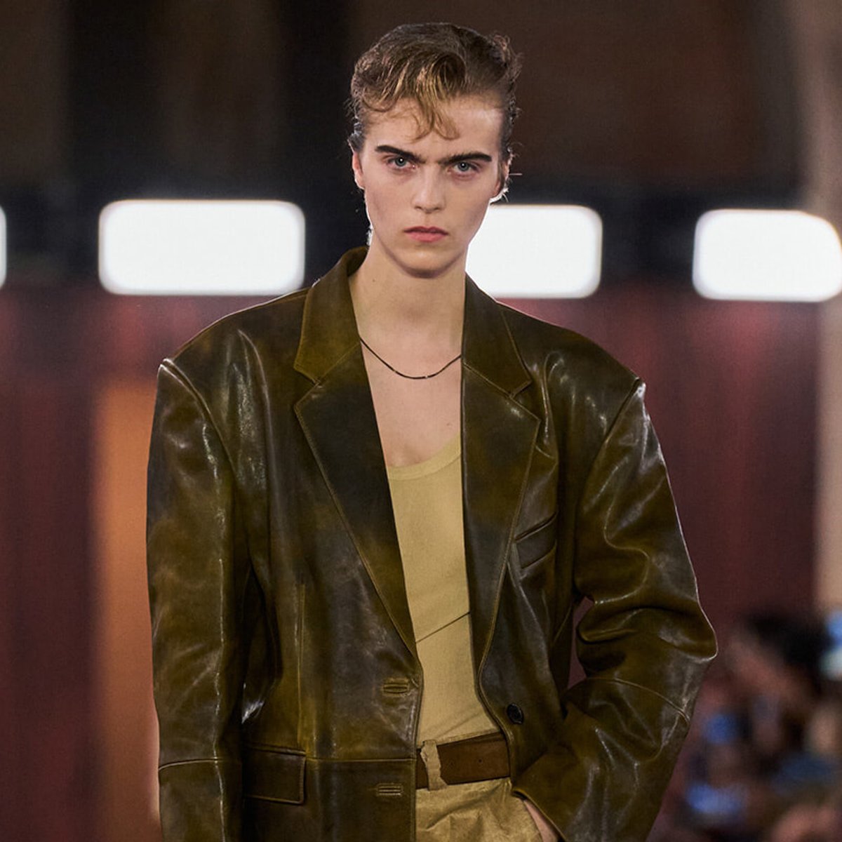 Model walking the runway in a dark brown leather blazer over a tan top, with a thin necklace and belt.