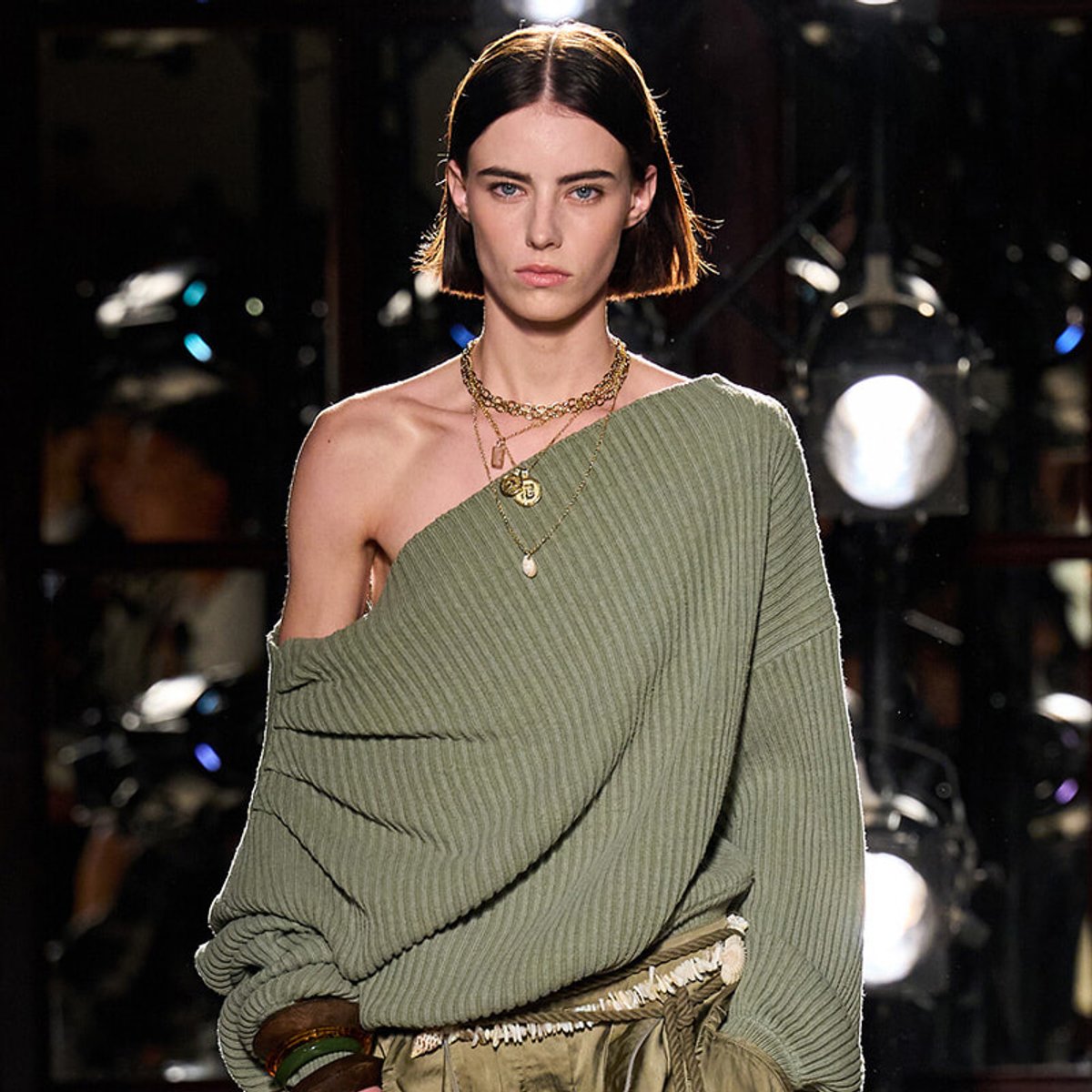 Model wearing an olive-green one-shoulder ribbed sweater on a runway, layered gold necklaces, short bob haircut.