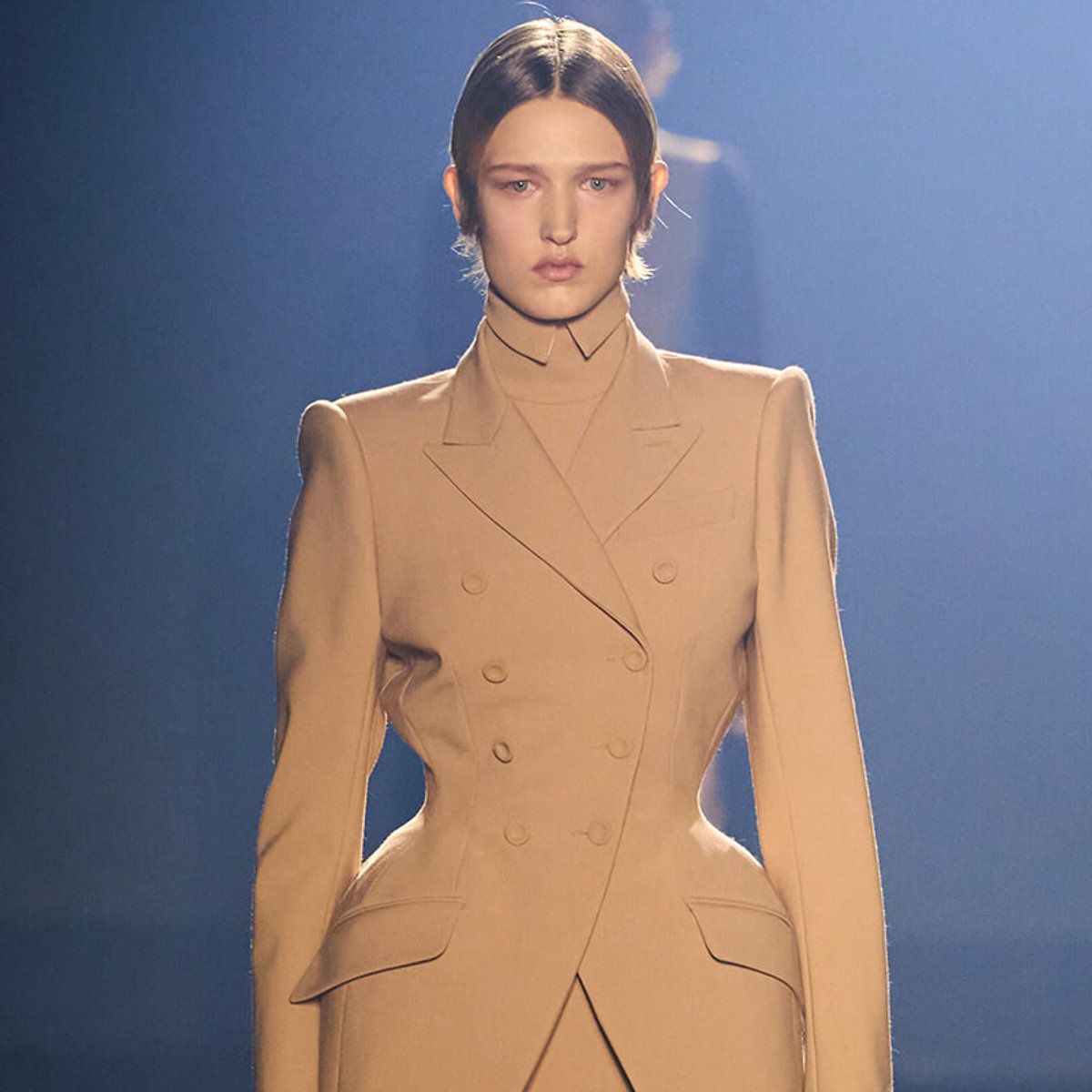 Model on a fashion runway wearing a tailored beige double-breasted blazer dress with structured shoulders and peak lapels under dramatic lighting.