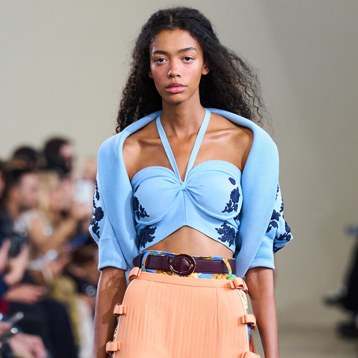 Model on a runway wearing a light blue cropped top with floral embroidery and a peach pleated skirt with a belt.