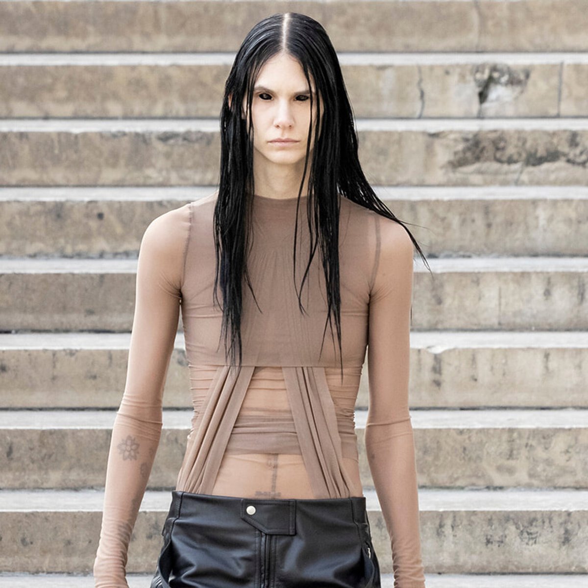 Model with long, wet black hair stands on concrete steps wearing a sheer beige top and black leather pants, looking straight ahead.